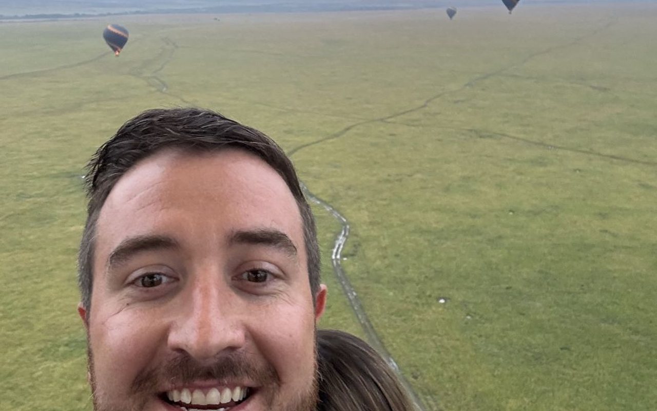 Andy smiling in a hot-air balloon basket at sunrise over the savanna, other balloons in the sky